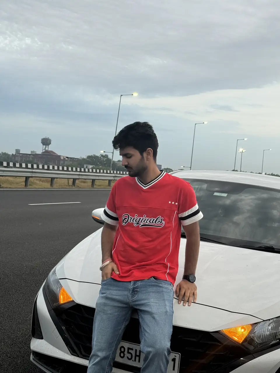 Pritish Bhatti in a red t-shirt leaning on a car, eyes down